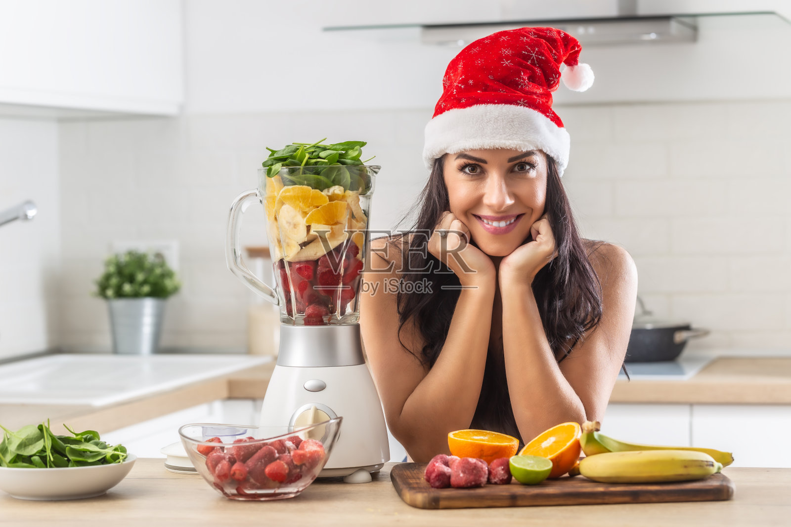 戴着圣诞帽的漂亮女人在厨房里，周围是水果和菠菜叶，用来做冰沙。照片摄影图片