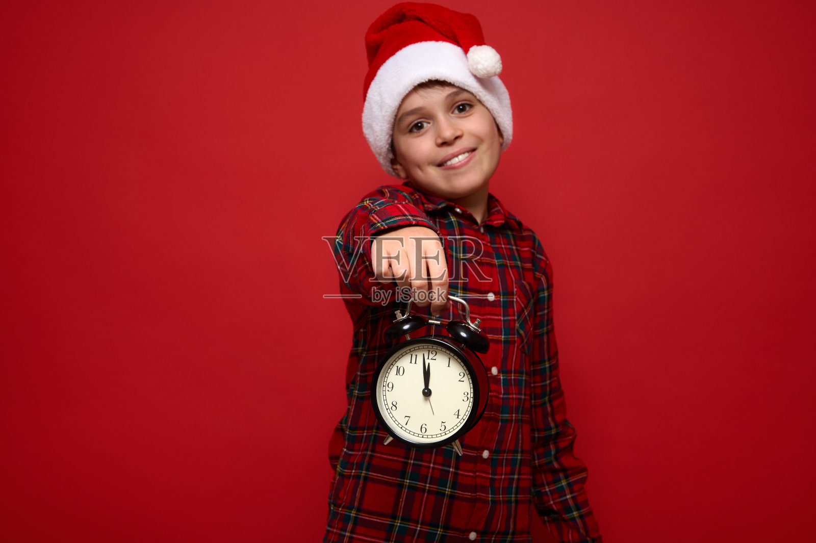 可爱的小男孩戴着圣诞老人的帽子，穿着红色格子衬衫，伸出双手，拿着一个黑色闹钟，闹钟上有午夜的刻度。圣诞节和新年的广告概念照片摄影图片