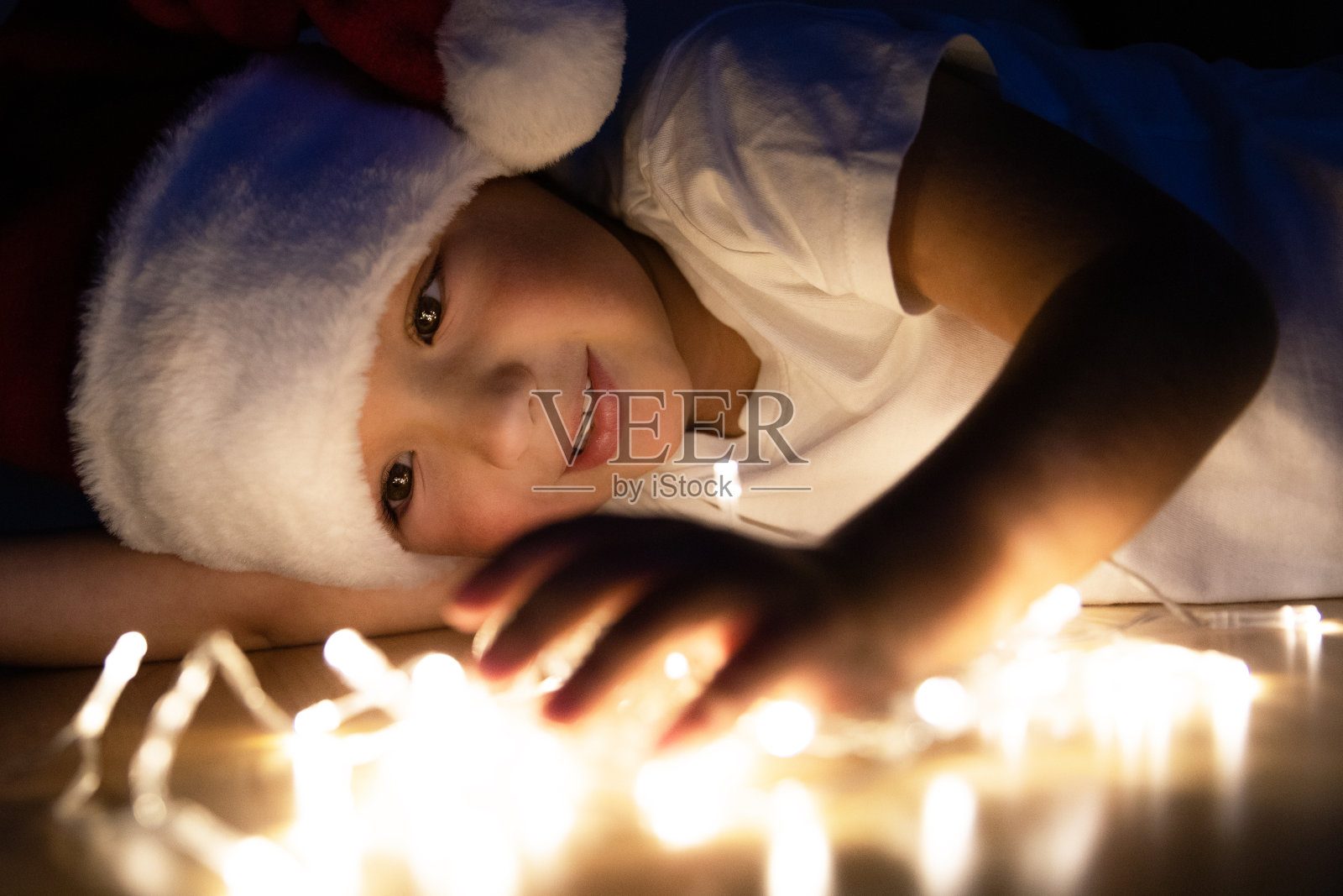 一个戴着圣诞帽、穿着白色t恤的小男孩在黑暗中躺在地板上玩圣诞灯。这孩子玩得很开心，想象力也很丰富。微笑着等待新年假期照片摄影图片