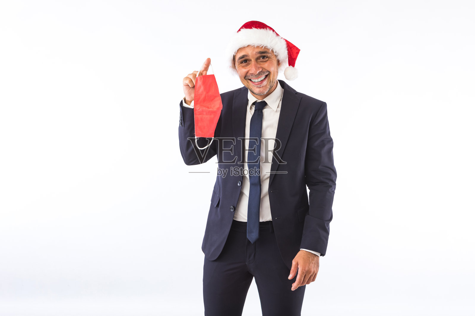 商人穿着西装，打着领带，戴着圣诞老人的圣诞帽，微笑着拿着红色的面具，在白色的背景下。圣诞节庆祝活动的概念。照片摄影图片