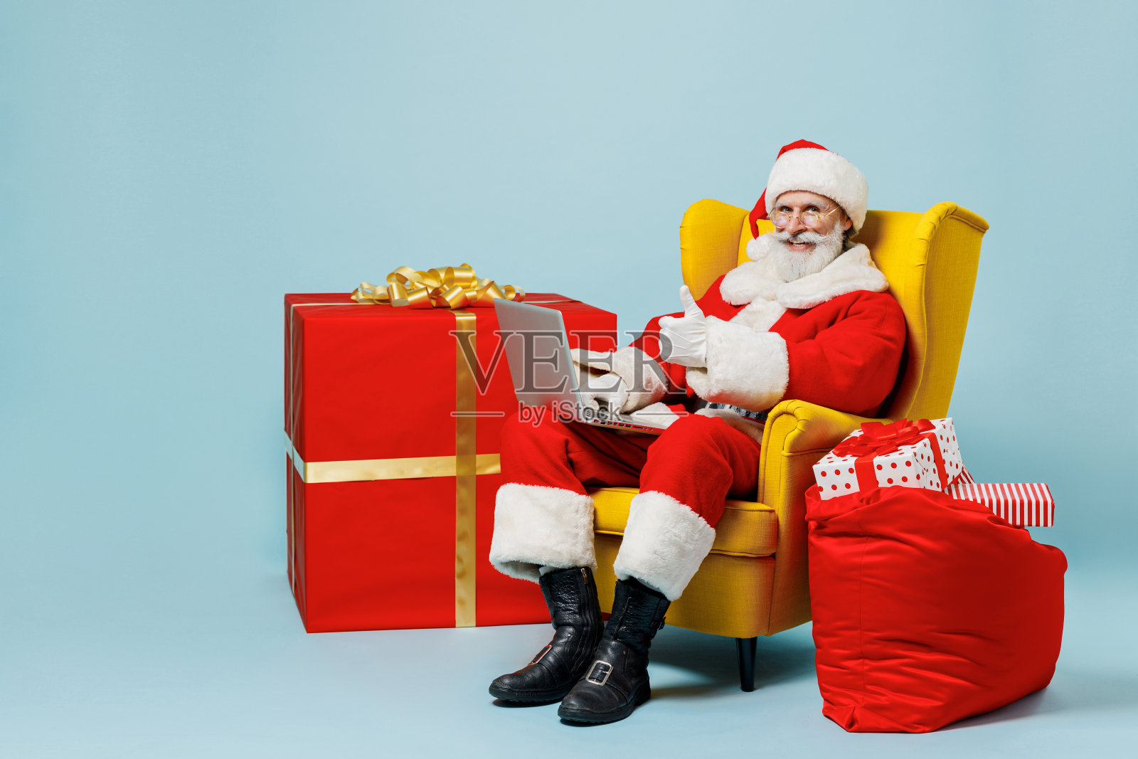 全身的圣诞老人戴着圣诞帽穿红色西装的人坐在椅子上用笔记本电脑电脑显示大拇指向上孤立在朴素的蓝色背景工作室新年快乐圣诞假期概念照片摄影图片