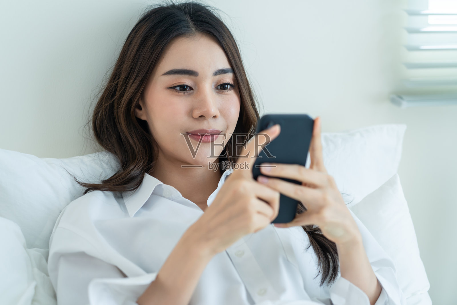 亚洲美女早上在家的床上用手机聊天。快乐休闲的美女躺在卧室里享受假日周末，在屋里用智能手机和技术交流。照片摄影图片