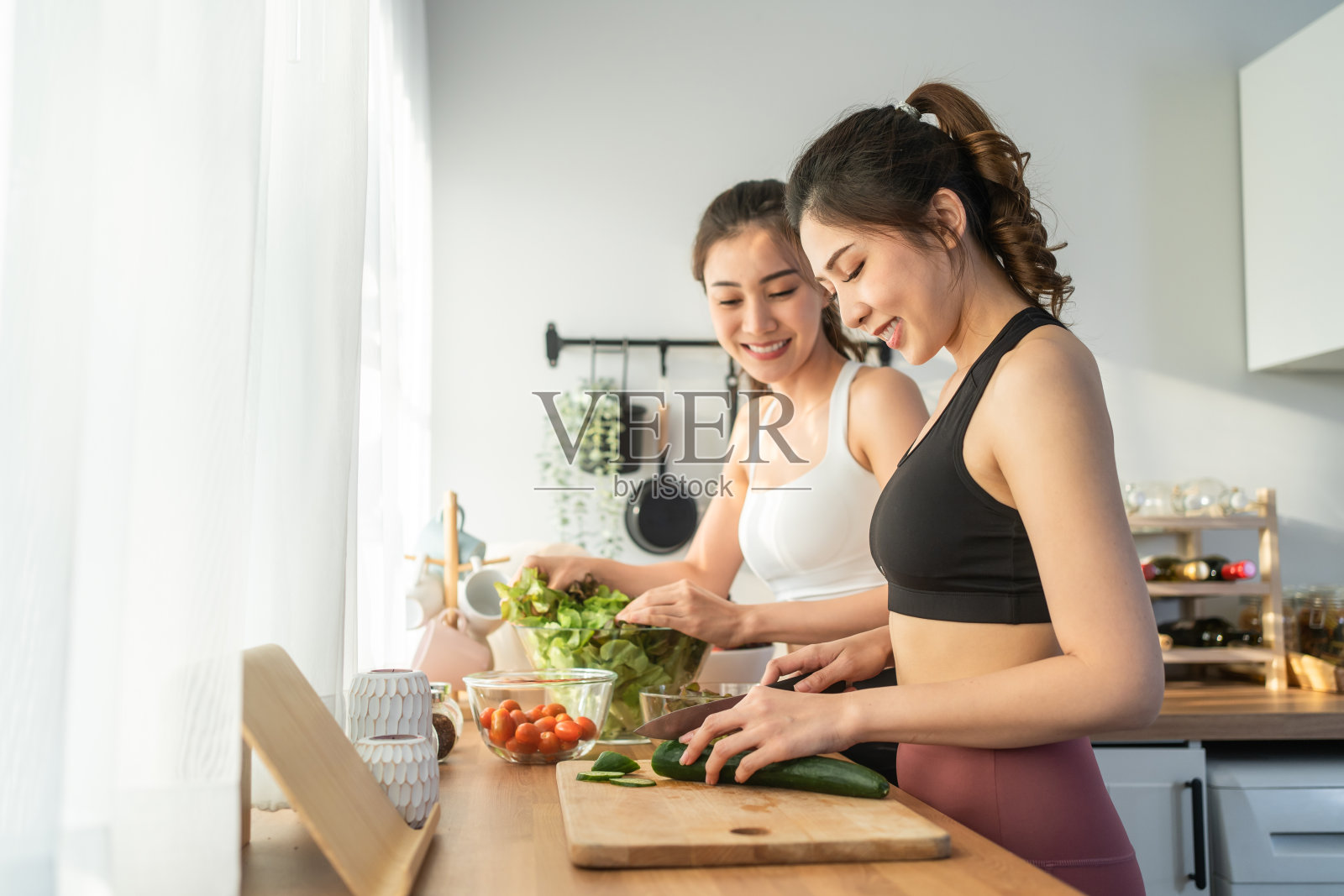 亚洲年轻的两个女子兄妹在厨房里穿着运动服烹饪沙拉。活泼漂亮的女孩妹妹心情愉快，喜欢吃蔬菜，健康饮食，减肥保健在家里照片摄影图片